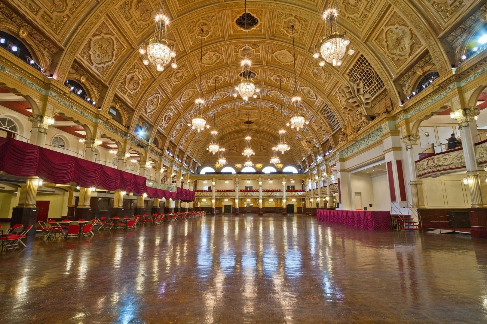 Ornate historic ballroom with chandeliers, red seating, and polished wooden floor